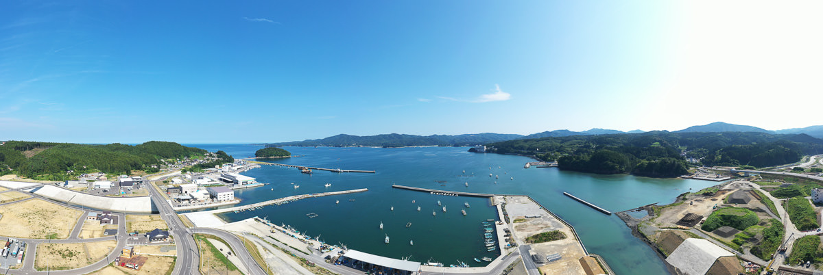 港に複数の船が停泊し、防波堤が海に向かって伸びている沿岸の町の広角風景で、町にはさまざまな建物や道路、緑地が広がり、背景には丘や山が見え、空は青く澄んでいる港町の写真