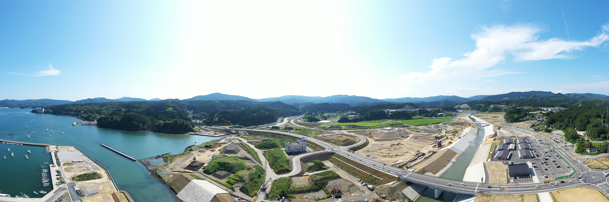 左側に海が広がり、岸辺には複数の船が停泊し、中央には川が流れて橋が架かり、右側には建物や道路などのインフラ整備が進む工事現場があり、背景には緑の丘や山が広がり、空は晴れて雲が少し浮かんでいる沿岸地域の写真