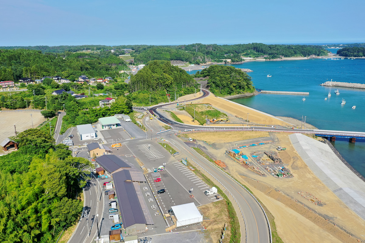 海岸に向かって道路が伸び、駐車場には複数の車が並び、建物や工事現場が点在し、海辺には砂地と船が停泊する水域があり、背景には緑豊かな丘と点在する家屋が見える沿岸地域の空撮写真