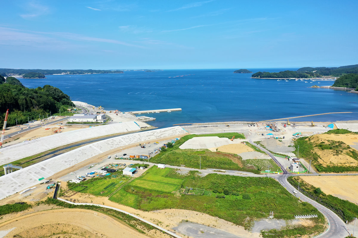 海に面した沿岸地域で、背景には広大な海が広がり、手前には開発中または整備中と思われる土地が区画ごとに分かれ、道路や小道が通り、緑地や草地、砂地のような場所も見られ、空は晴れて雲が少ない穏やかな天気の写真