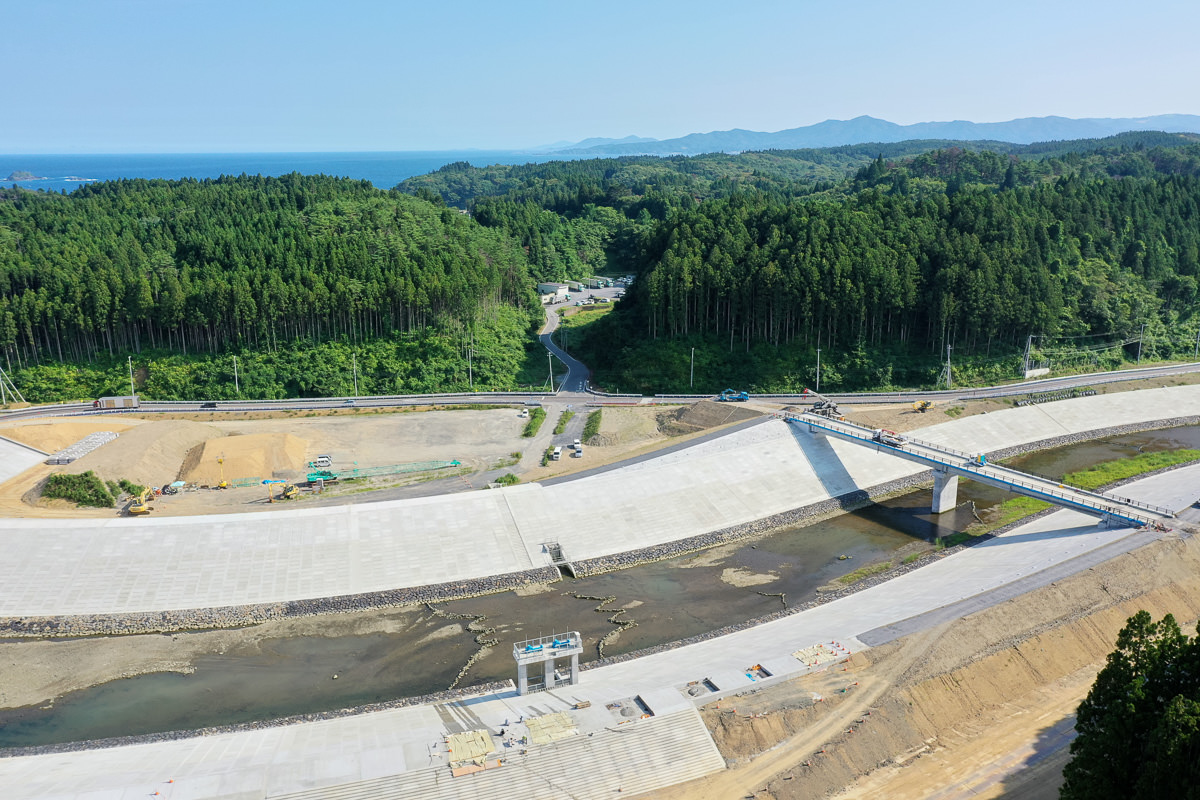 森林に囲まれた地域にある大きなダムの構造物で、コンクリートの壁とその上を通る橋があり、ダムへと続く道路や工事用の機材が見られ、背景には木々と丘が広がり、空は晴れているダム施設の写真
