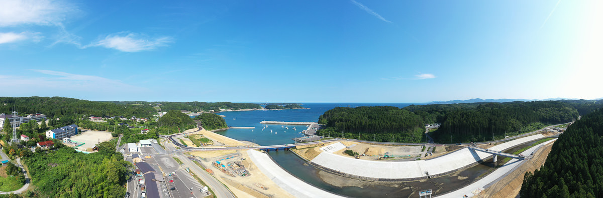 緑豊かな森林に囲まれた沿岸地域の広角風景で、川が海へと流れ込み、複数の橋が川を横断し、左側には建物や道路が広がり、右側には密集した森林が広がっていて、空は晴れて雲がほとんどない自然と人の営みが調和した写真