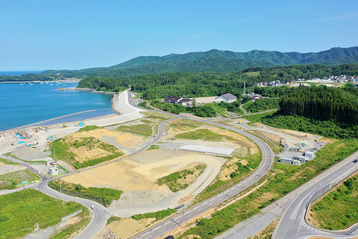 海に面した沿岸地域の空撮で、左側に海が広がり、海岸線には開発中の区域があり、陸地には曲がりくねった道路や緑地、建設中または整備された区画が点在し、背景には森林に覆われた丘や山が広がり、建物も散在して人の生活やインフラの存在が感じられる晴天の写真