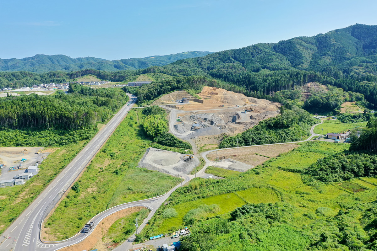 緑の丘や森林に囲まれた風景の空撮で、中央には道路が通り、一部の土地は整地や掘削がされていて建設または採掘のための作業が行われている可能性があり、空は青く晴れている写真