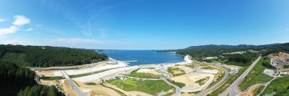 海に面した沿岸地域の広角風景で、背景には広大な海が広がり、海岸線には砂浜と人工の防波堤があり、手前には道路や緑の畑、開発中と思われる整地された土地が広がり、周囲には森林や丘陵が広がっていて、空は晴れて雲がほとんどない写真