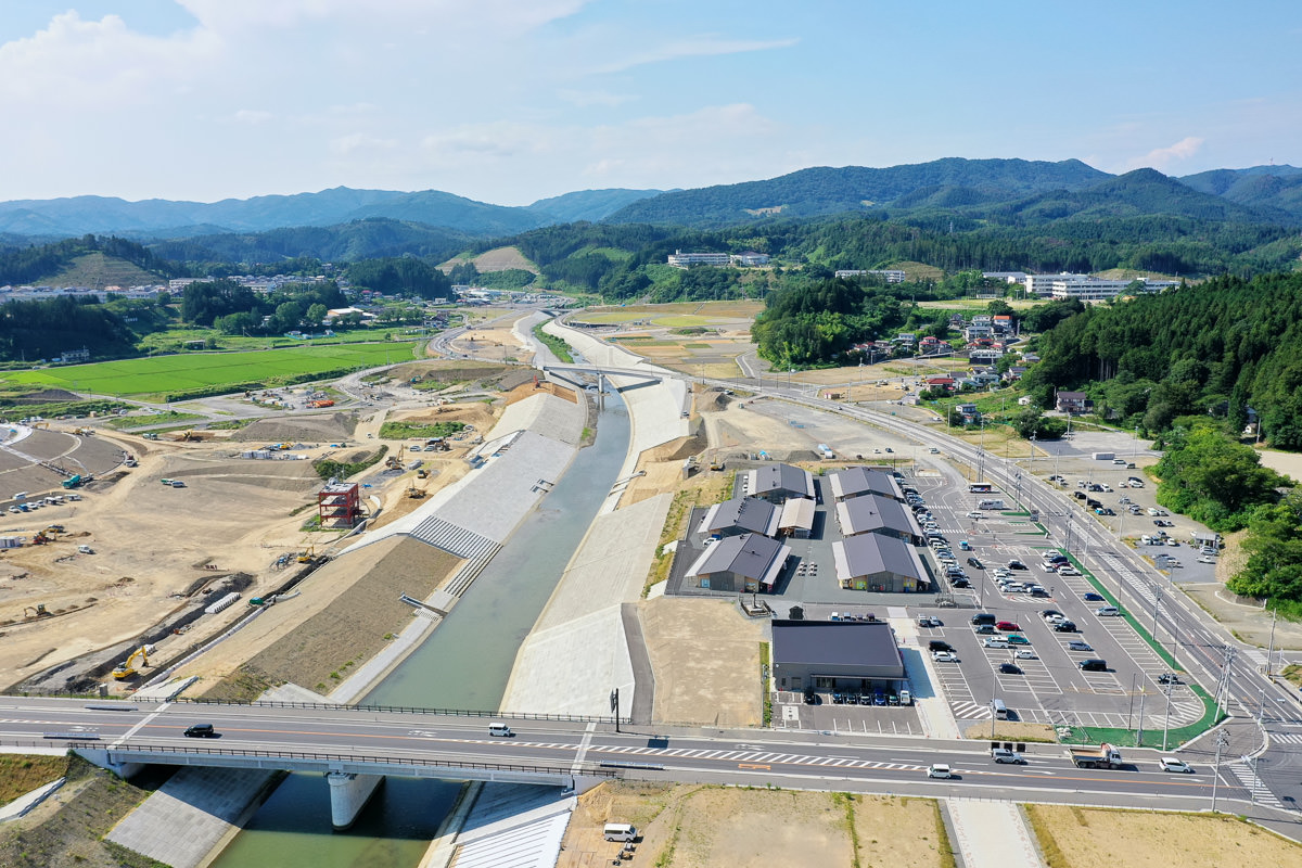 中央に川が流れる農村地域の空撮で、手前には川に架かる橋があり、右側には複数の建物と駐車場が見られ、周囲には畑や丘、森林が広がっている自然と人の営みが調和した風景の写真