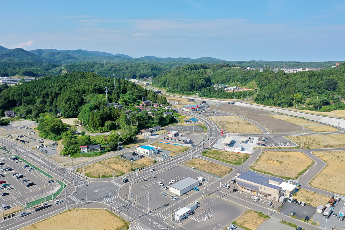 森林に覆われた丘と整備された土地が混在する農村地域の空撮で、複数の建物や駐車場、道路が見られ、緑の木々や開けた畑が広がり、道路には車が整然と駐車されていて、背景には緑豊かな丘陵が広がり、空は青く澄んでいる写真