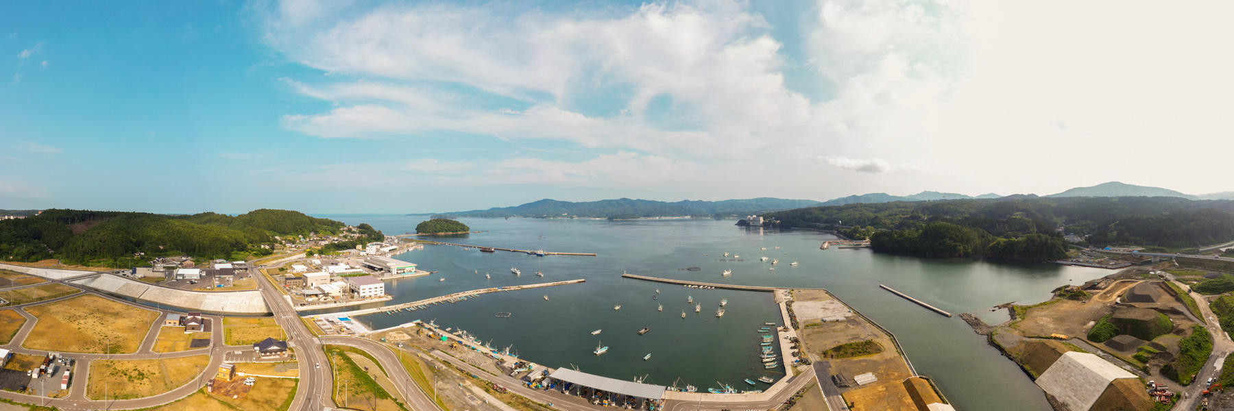 緑の丘と山々に囲まれた海辺の町を広く見渡す風景で、港には複数の船が停泊しており、町の中心には建物や道路が整然と広がっている様子が見える写真
