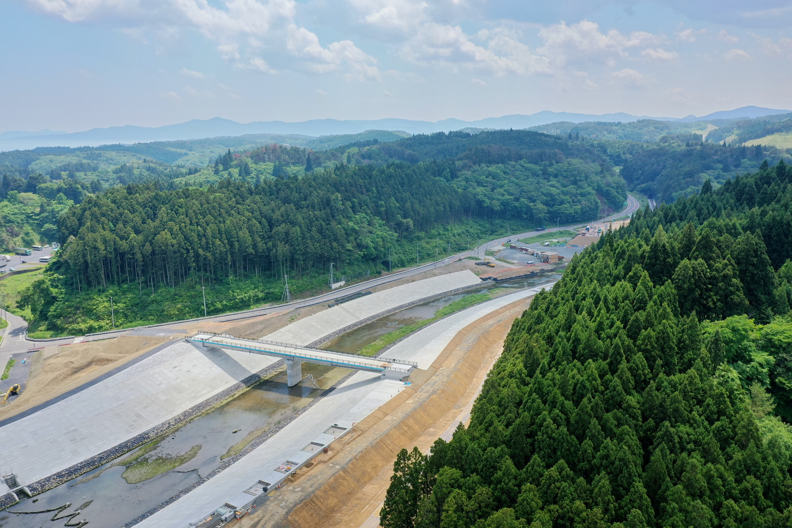 川または運河に架かる新しく建設された橋を中心に、両側に広がる谷をつなぐ形で構造物が配置され、周囲には密集した森林と丘陵地が広がり、洪水対策や交通インフラの一環と思われる工事現場を上空から撮影した橋梁工事の写真