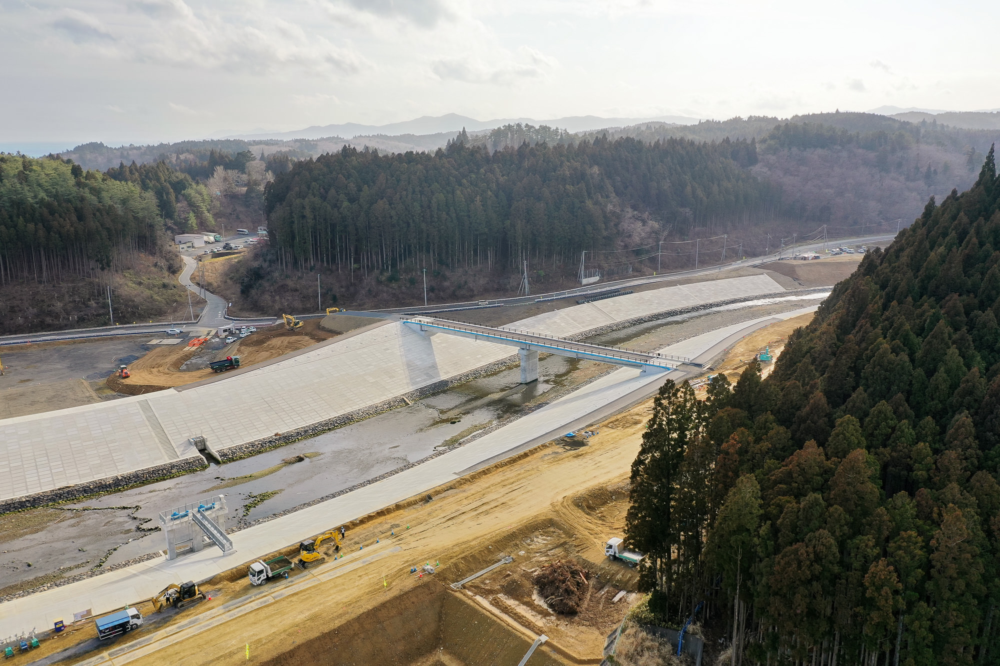 川または運河に架けられた新しい橋を中心とした工事現場を空から撮影した写真で、周囲には森林と丘が広がり、建設車両や機材が点在する田舎または郊外の風景が見える写真