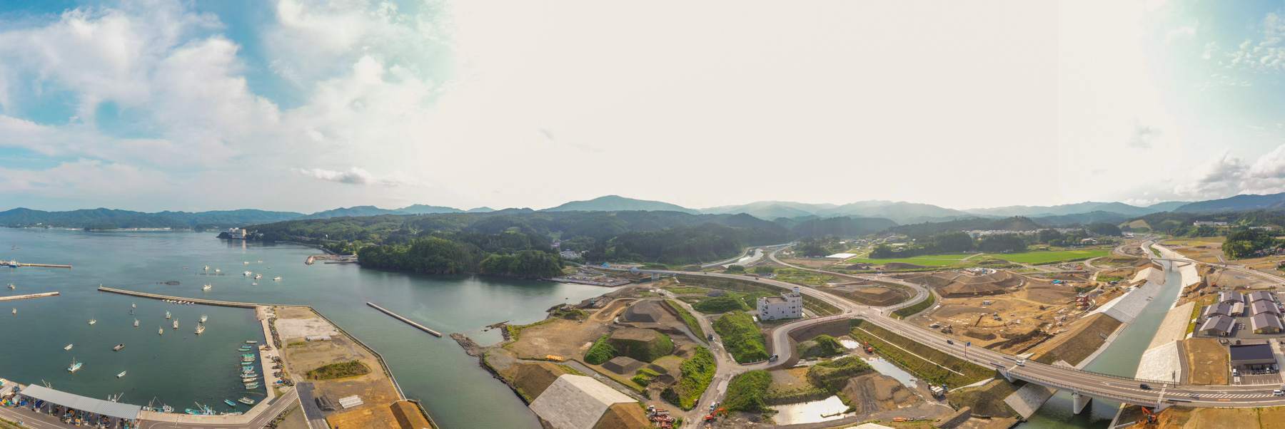 左側に港があり複数の船が停泊している海辺の地域を広く見渡す風景で、町には建物や道路、橋などのインフラが整備され、背景には緑の丘や山々が広がっている写真