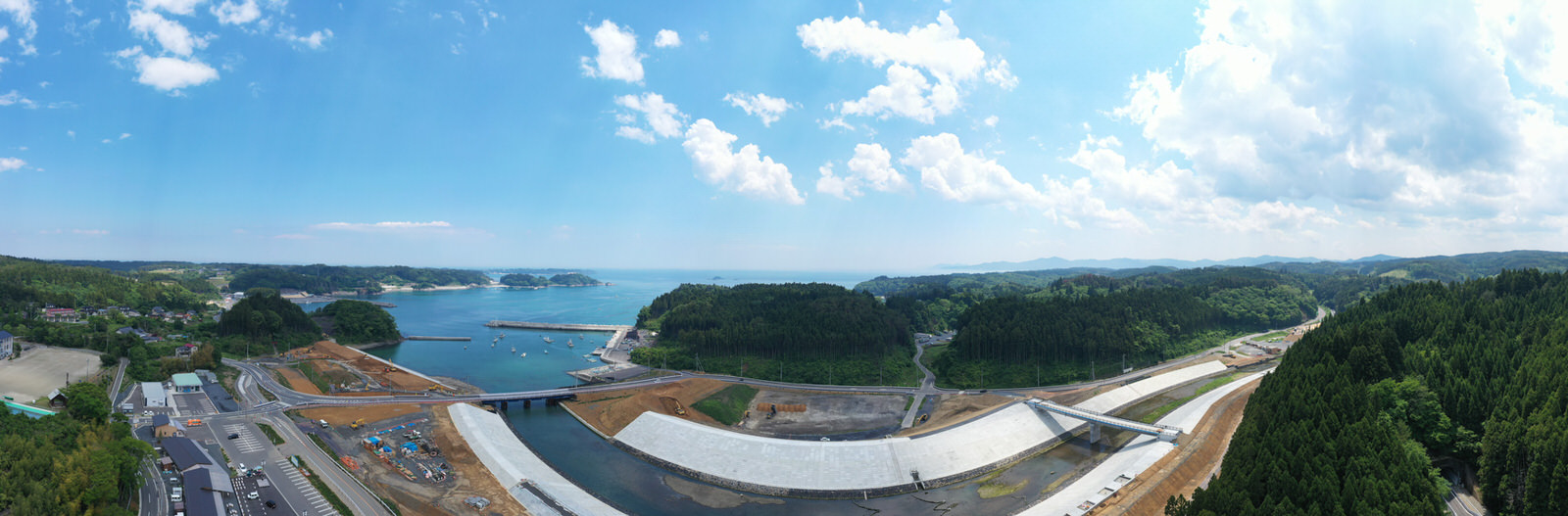 広大な海を背景に、手前には防潮堤と思われる大規模な建設現場が広がり、周囲には緑豊かな森林と丘があり、複数の道路や建物が点在し、空には雲が浮かぶ海沿いのインフラ工事の写真