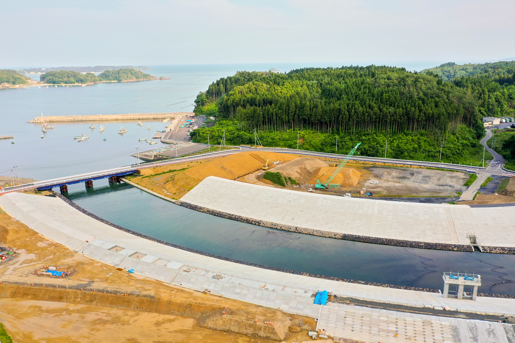 海沿いにある大きなコンクリート構造物が水辺に隣接し、橋がその上を横断していて、港には複数の船が停泊し、背景には緑豊かな森林と丘が広がっている写真