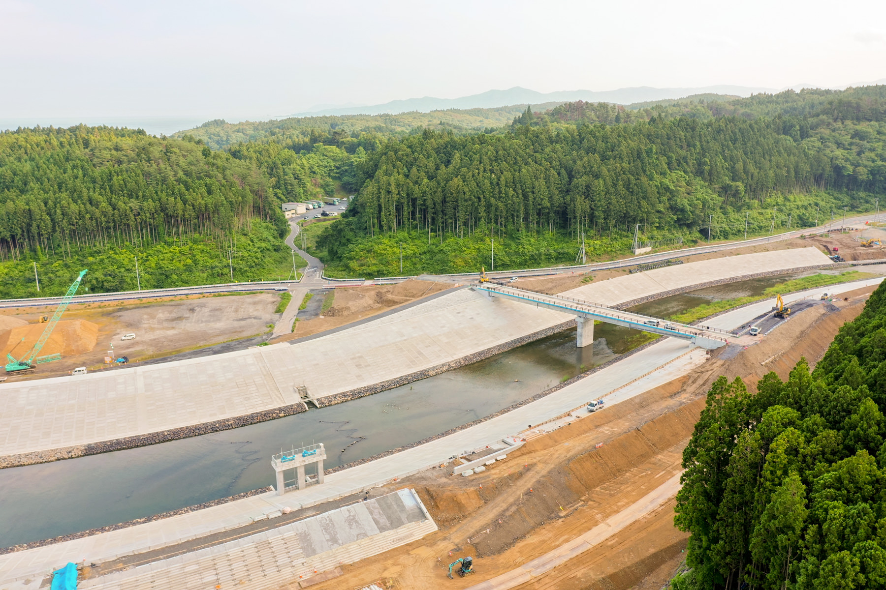 中央に川が流れるコンクリート構造の大規模な工事現場が広がり、橋がその上を横断し、周囲には密集した森林と丘陵が広がっていて、現場には建設機械が点在している写真