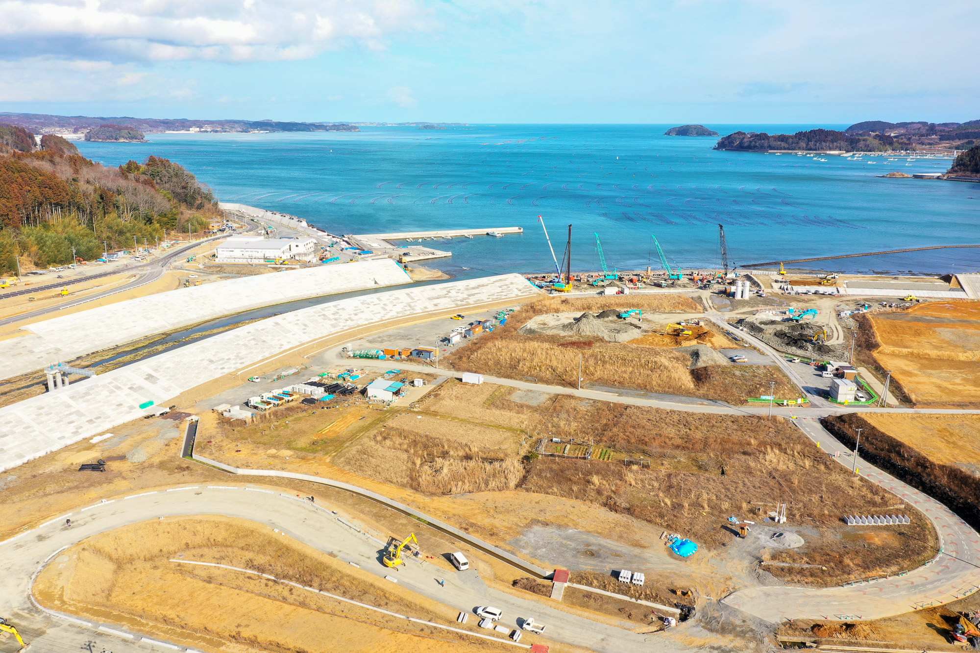 海岸沿いの工事現場で複数のクレーンや建設機械が稼働しており、背景には青く広がる海と点在する島々が見える写真