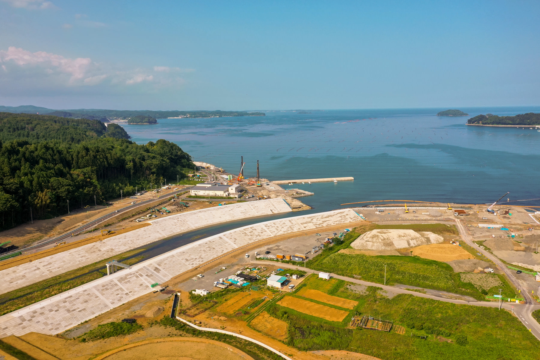 海岸沿いに広がる大規模な工事現場で、複数のクレーンや建設機材、建物や構造物が見られ、周囲には緑豊かな森林が広がり、海は遠くの小島まで続き、空には少し雲が浮かんでいる沿岸開発地域の写真