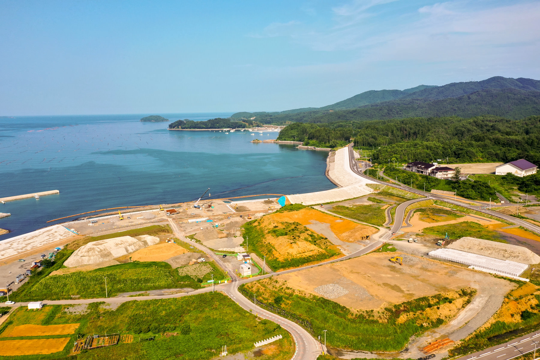 海岸沿いに道路が通り、さまざまな色の区画が広がっていて、開発の進行状況や植生の違いが見られ、道路の先には小さな港があり、海は穏やかで、背景には緑に覆われた丘が広がり、空には薄い雲が浮かんでいる沿岸地域の写真