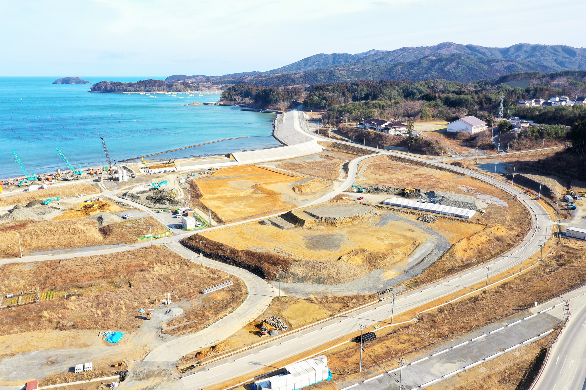 海岸沿いの工事現場で複数の区画が開発されており、道路や小道が敷地内を曲がりくねって走り、海岸近くには建設機材が見え、岩場と砂浜が混在する海岸線の奥には広がる海と遠くの丘や森林が見える写真
