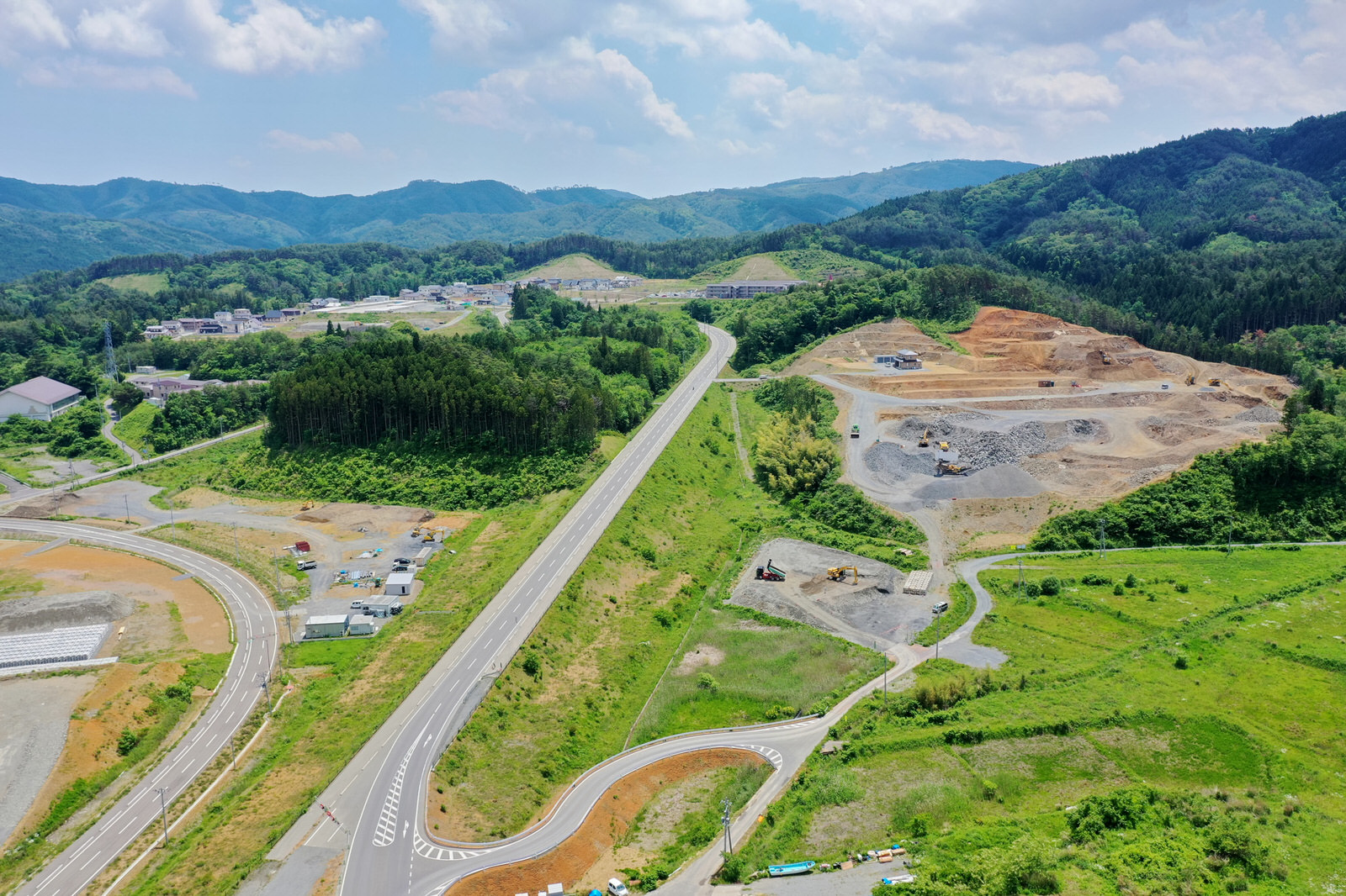 中央に道路が通る風景を上空から撮影した写真で、左側には曲がりくねった道路と小さな建物が点在し、右側には土砂や砂利が積まれた工事中または掘削中の土地が広がり、背景には緑豊かな丘や山々が見え、空には雲が浮かぶ造成地の写真