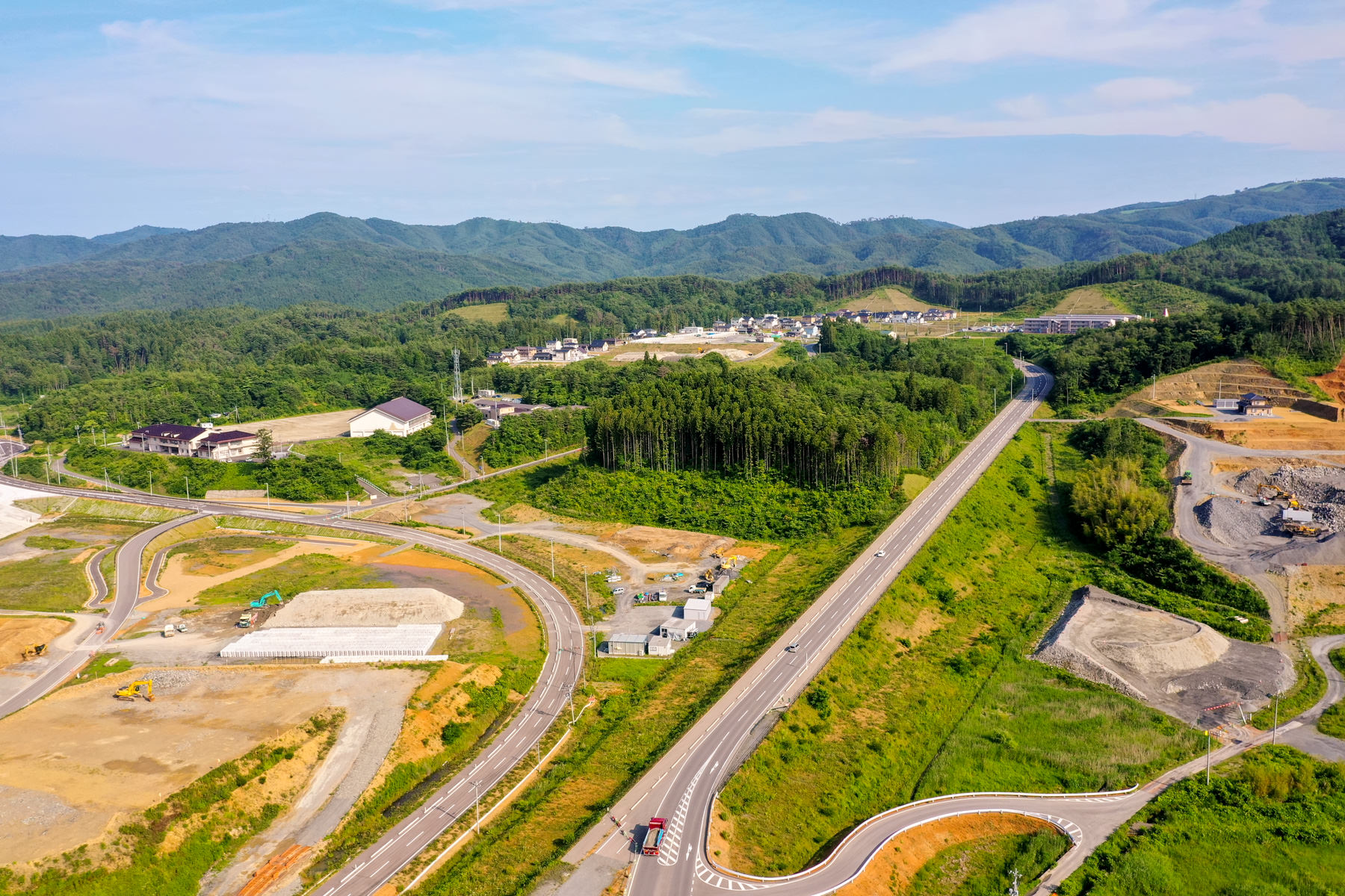 曲がりくねった道路と直線の道路が緑豊かな風景の中を通り、所々に工事区域や建物が点在し、背景には木々に覆われた丘が広がっている農村地域の空撮写真