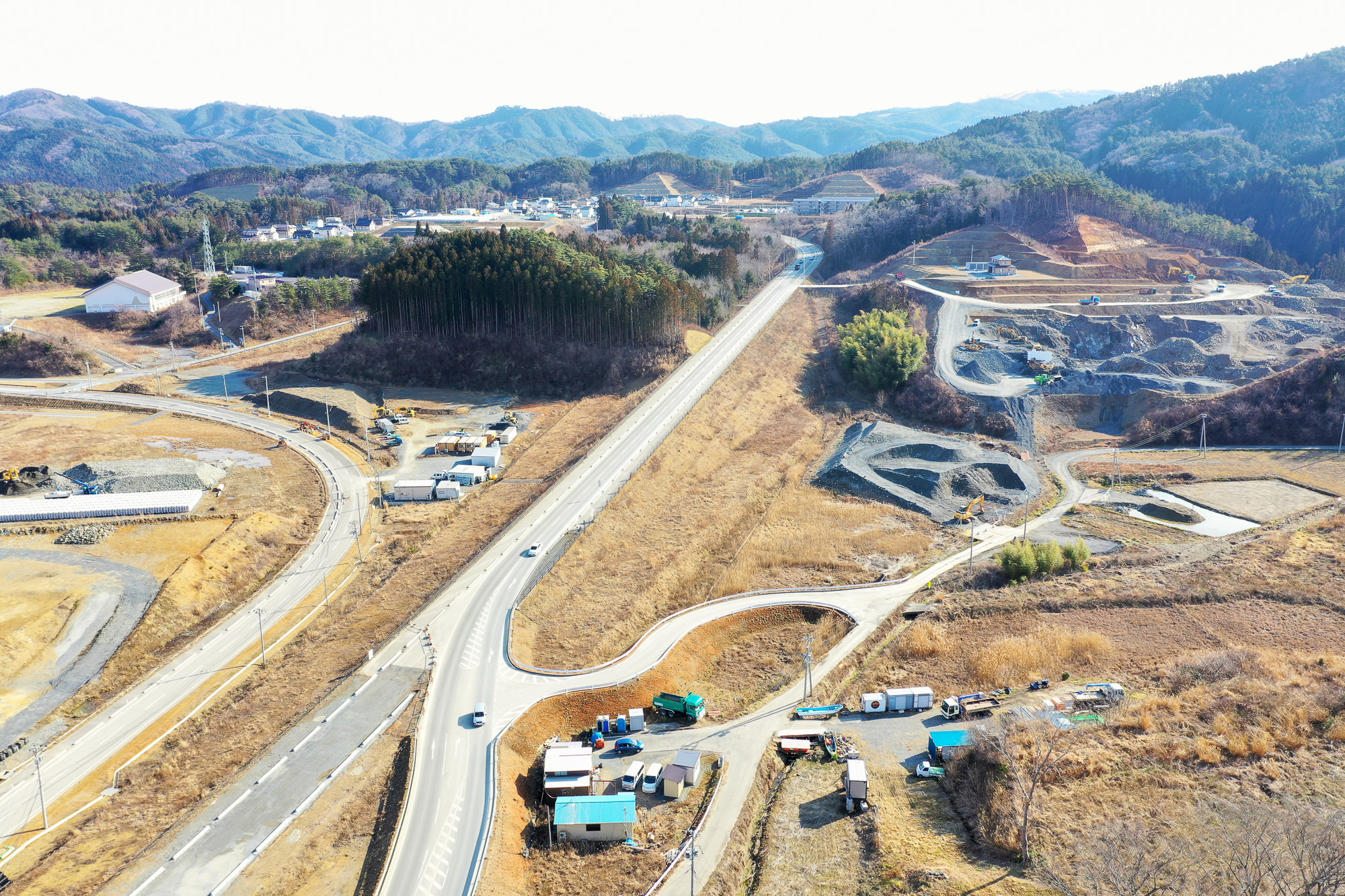 田園地帯を空から見下ろした写真で、中央には道路が通り、そこから複数の小道が分岐し、周囲には建物や構造物が点在し、丘陵地には森林と開けた畑が広がり、背景には木々に覆われた山々が見える写真