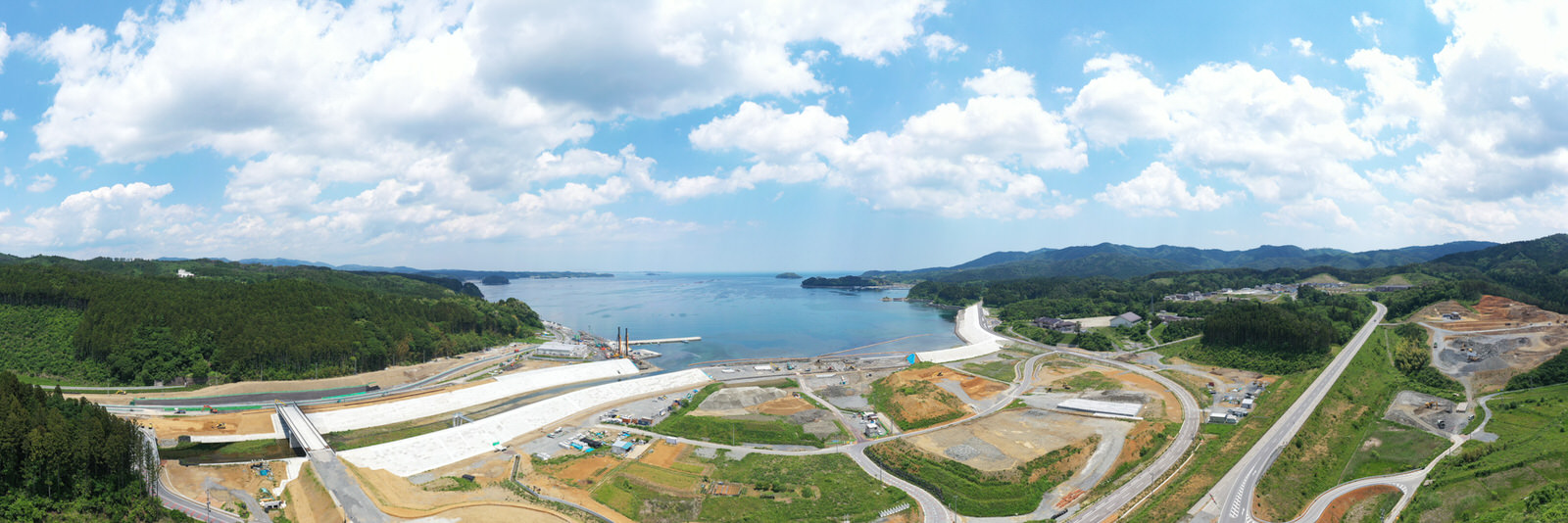 海に面した地域を広く見渡す写真で、道路や整地された土地、そして水辺近くには大きなコンクリート構造物があり、背景には水平線まで広がる水面と森林に覆われた丘が見え、空には雲が点在する海辺の開発地域の写真