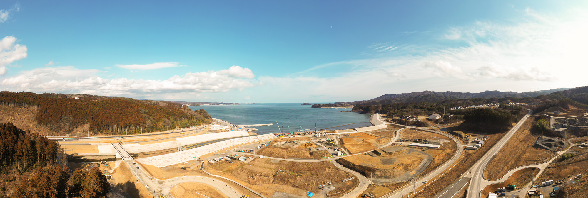 開発中の海岸地域を広く見渡した写真で、中央には複数の道路や造成地が広がり、一部は整地や掘削が行われており、背景には海岸線と水平線まで続く海が見え、両側には丘や森林が広がる写真
