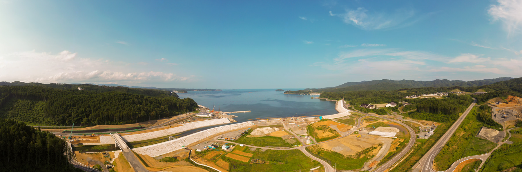 広大な海に面した沿岸地域の広角風景で、周囲には緑の丘や森林が広がり、複数の道路や工事現場が見られてインフラ整備が進行中であり、空は晴れて雲が少し浮かんでいる開発中の海辺の写真