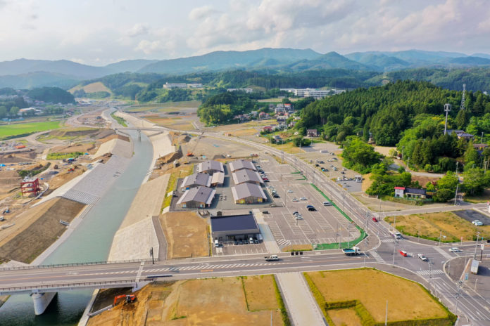 川が流れる農村地域の空撮で、川には橋が架かり、片側には複数の建物と駐車場があり、背景には丘や山が広がり、周囲には緑と木々が点在し、空には雲が少し浮かんでいる自然と人の営みが見える風景の写真