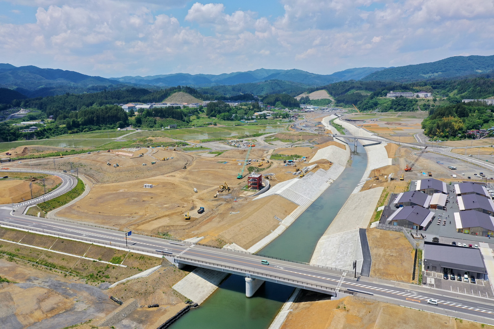 中央に川が流れ、その上に橋が架かり、両岸には工事車両や機材が点在する建設現場を上空から撮影した写真で、周囲には道路や建物があり、背景には緑豊かな丘が広がり、空には雲が浮かぶ河川工事の写真