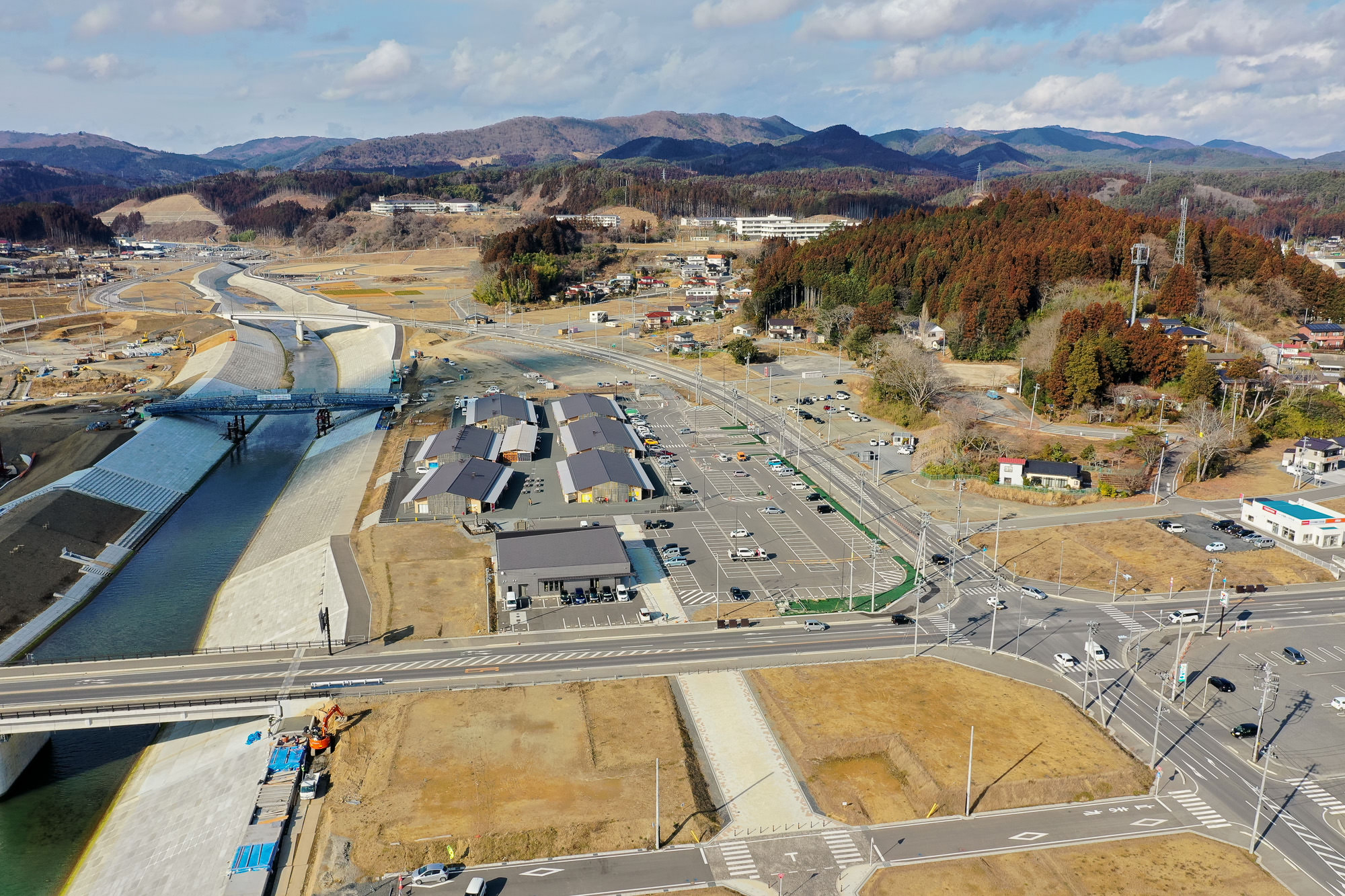 川が流れる田園地帯を空から撮影した写真で、複数の建物や道路、駐車場が点在し、背景には木々に覆われた丘や山々が広がる写真