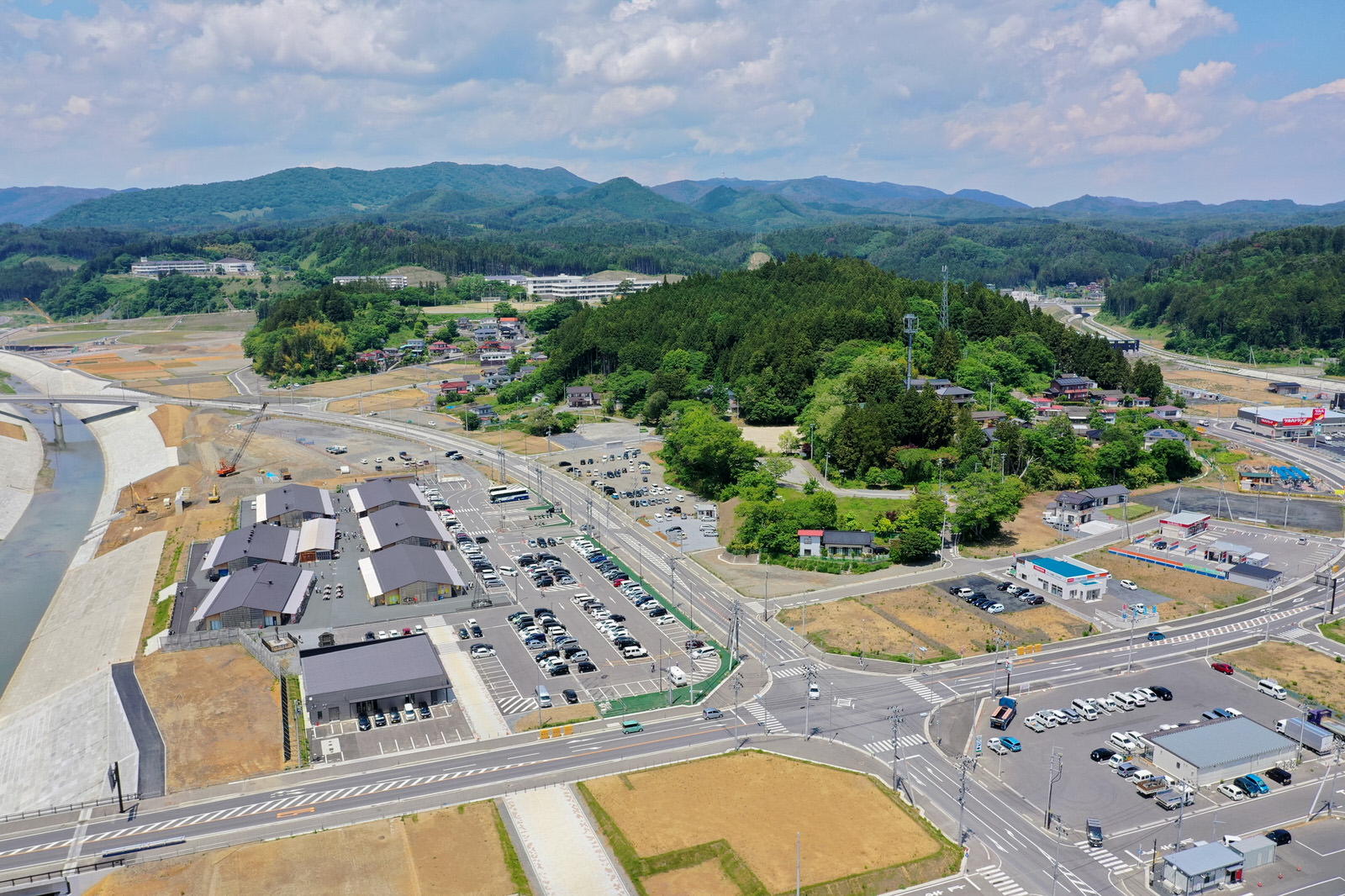 緑豊かな丘や山々に囲まれた小さな町または村を上空から撮影した写真で、複数の建物や駐車場、交差点を含む道路網が整然と広がり、左側には堤防に囲まれた川が流れる地域のインフラが整った町の写真