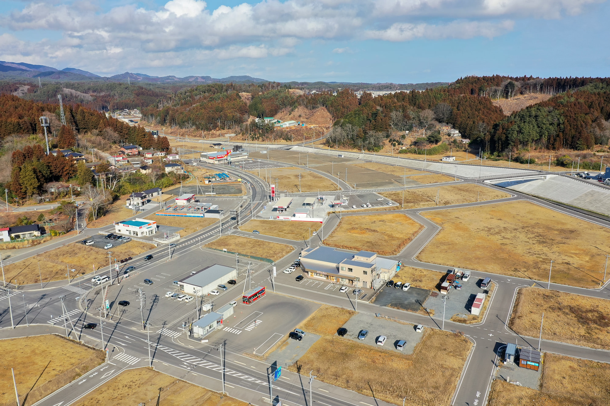 田園地帯を空から撮影した写真で、複数の建物や道路、駐車場が点在し、草地や木々が広がる中に交差点や車両が見え、背景には丘があり、空には雲が少し浮かぶ写真