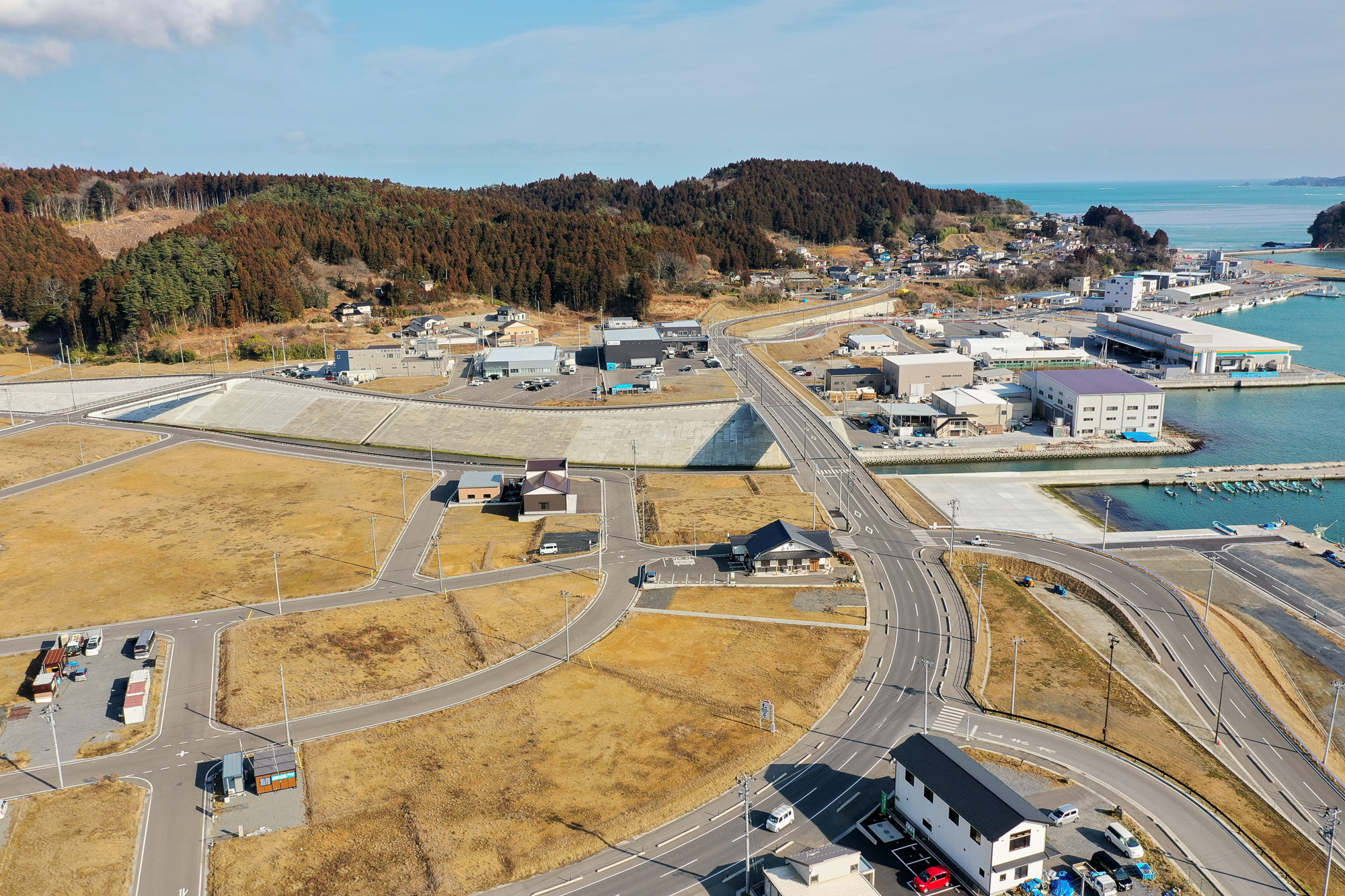 海沿いの町を空から撮影した写真で、複数の建物や道路、港が広がり、道路や駐車場には車両が見え、周囲には草地や木々のある丘が背景にあり、右側には海が広がる写真