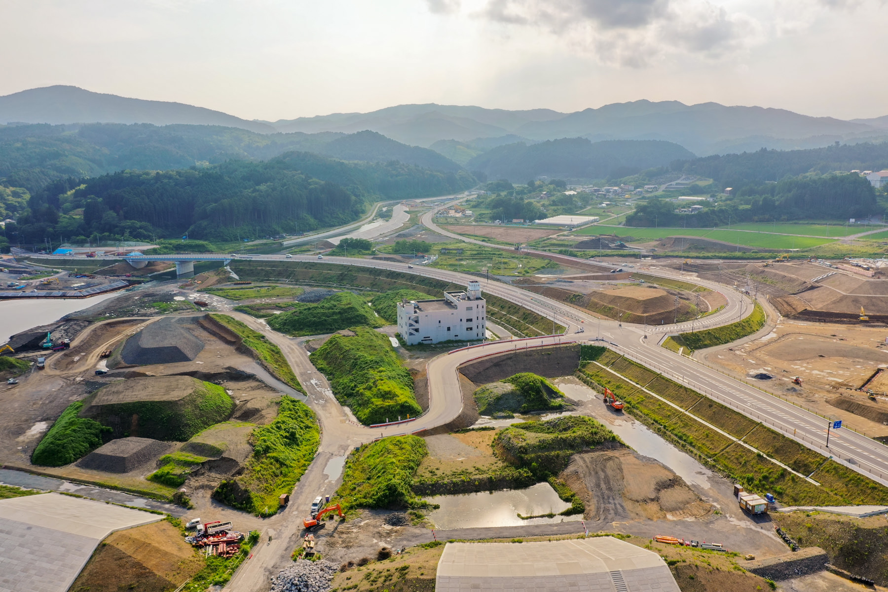 中央に白い建物があり、その周囲に緑地や土の盛り上がりが広がる工事現場を上空から見下ろした風景で、複数の道路や通路が整備されつつあり、建設機械が点在し、背景には丘や山々が広がっている写真