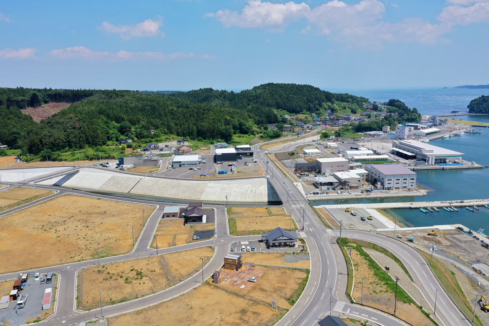 海沿いの町を上空から撮影した写真で、道路や建物、港が広がり、倉庫や住宅などの構造物が点在し、右側には海が広がって船が停泊している港があり、背景には緑の丘が広がる港町の写真