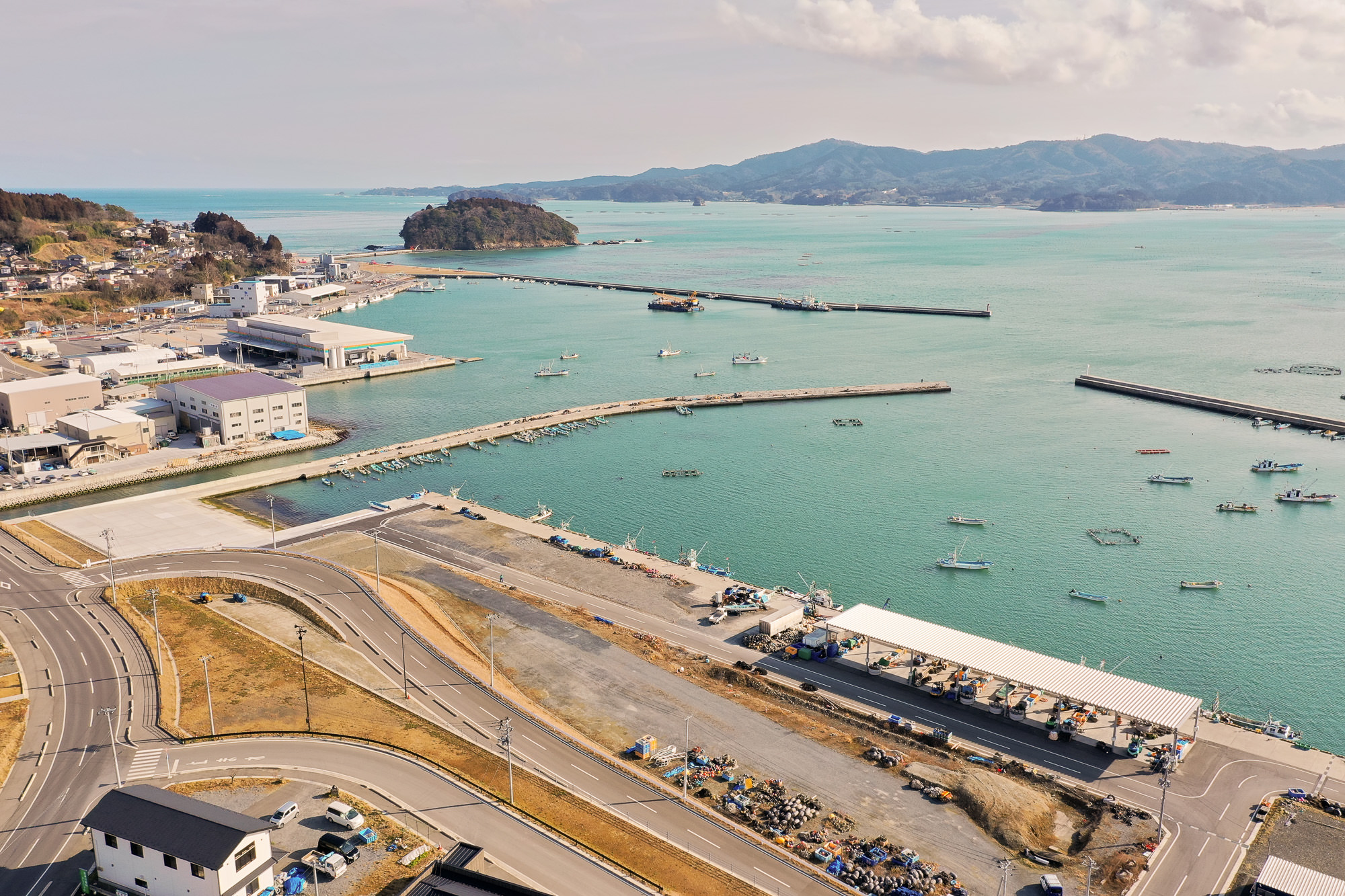 港のある海沿いの町を空から撮影した写真で、複数の桟橋が海に突き出し、小型船が多数停泊しており、倉庫や住宅などの建物が海岸近くに並び、手前には道路網と駐車中の車両が見え、背景には湾を囲む丘や山々が広がる写真