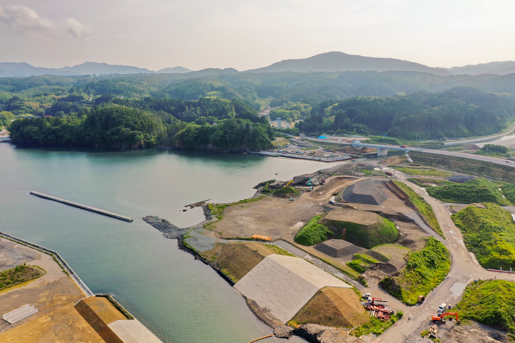 海に面した地域を上空から見下ろした風景で、複数の工事現場に重機や資材が並び、周囲には緑の丘や山々が広がり、空には雲が点在している写真