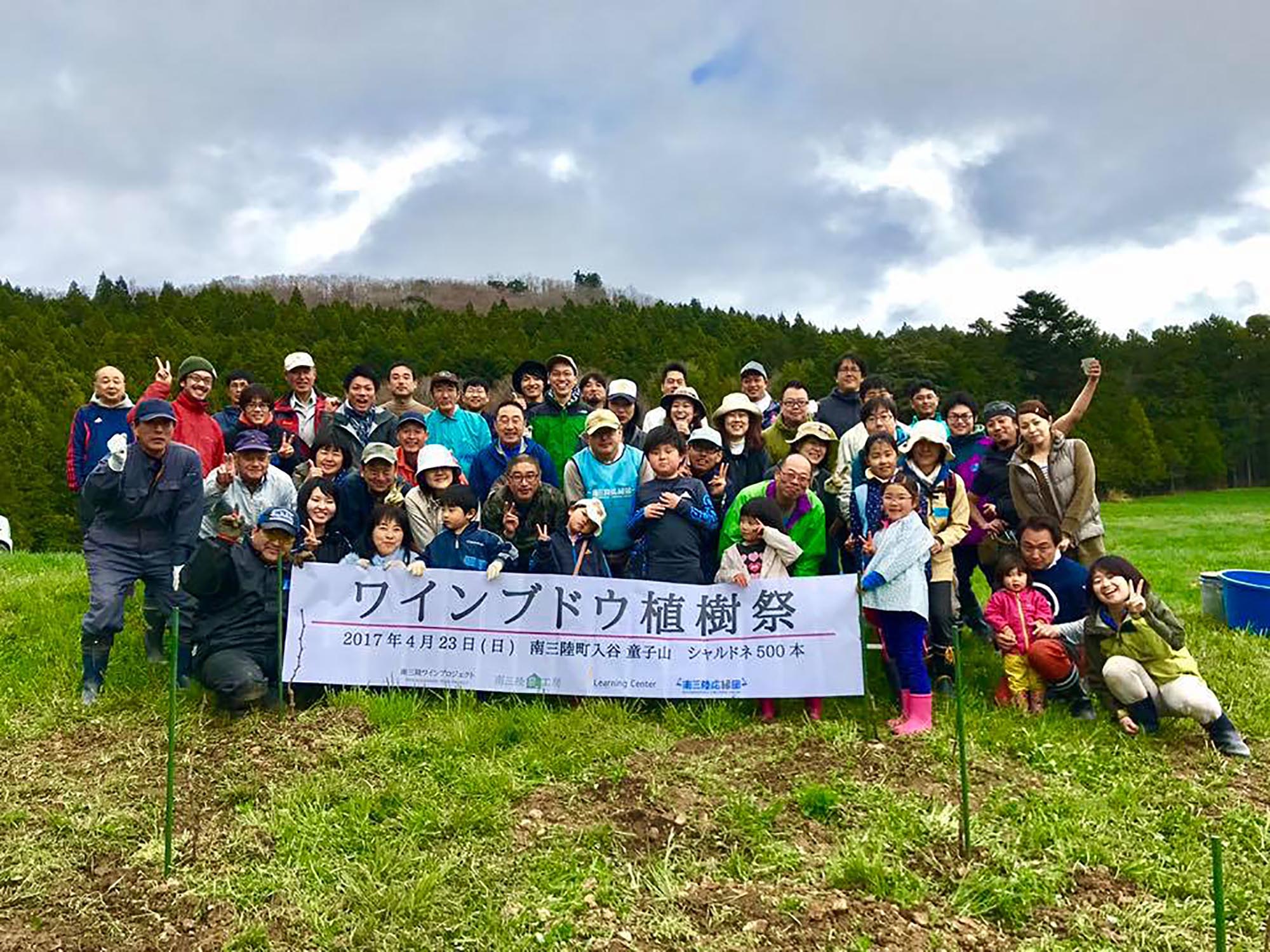 「ワインブドウ植樹祭」の横断幕を掲げ、老若男女の多くの参加者が笑顔で記念撮影している集合写真
