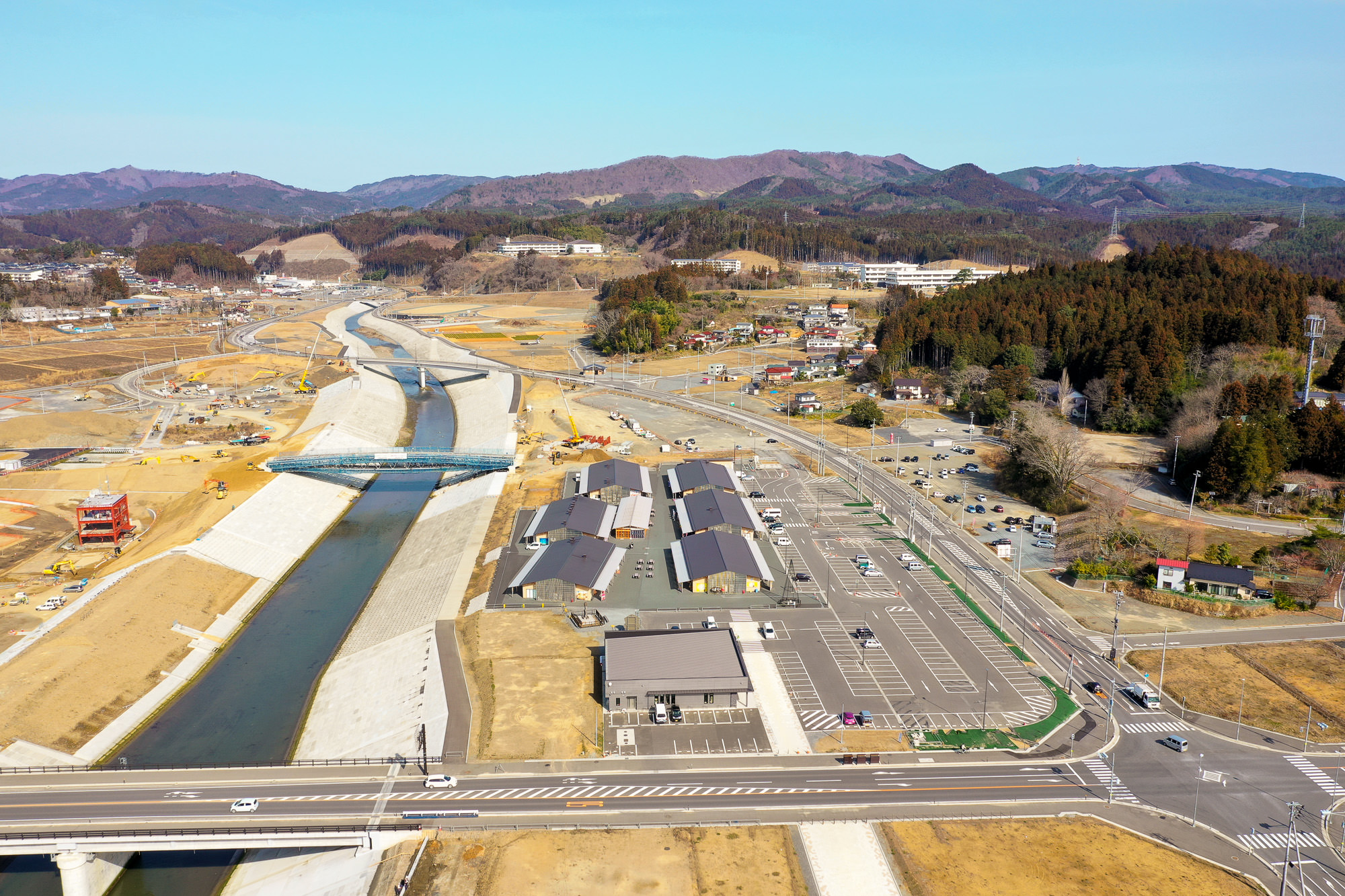 広々とした駐車場の奥に川が流れ、その周囲には緑豊かな田舎の風景が広がっている様子を写した田舎の風景の写真