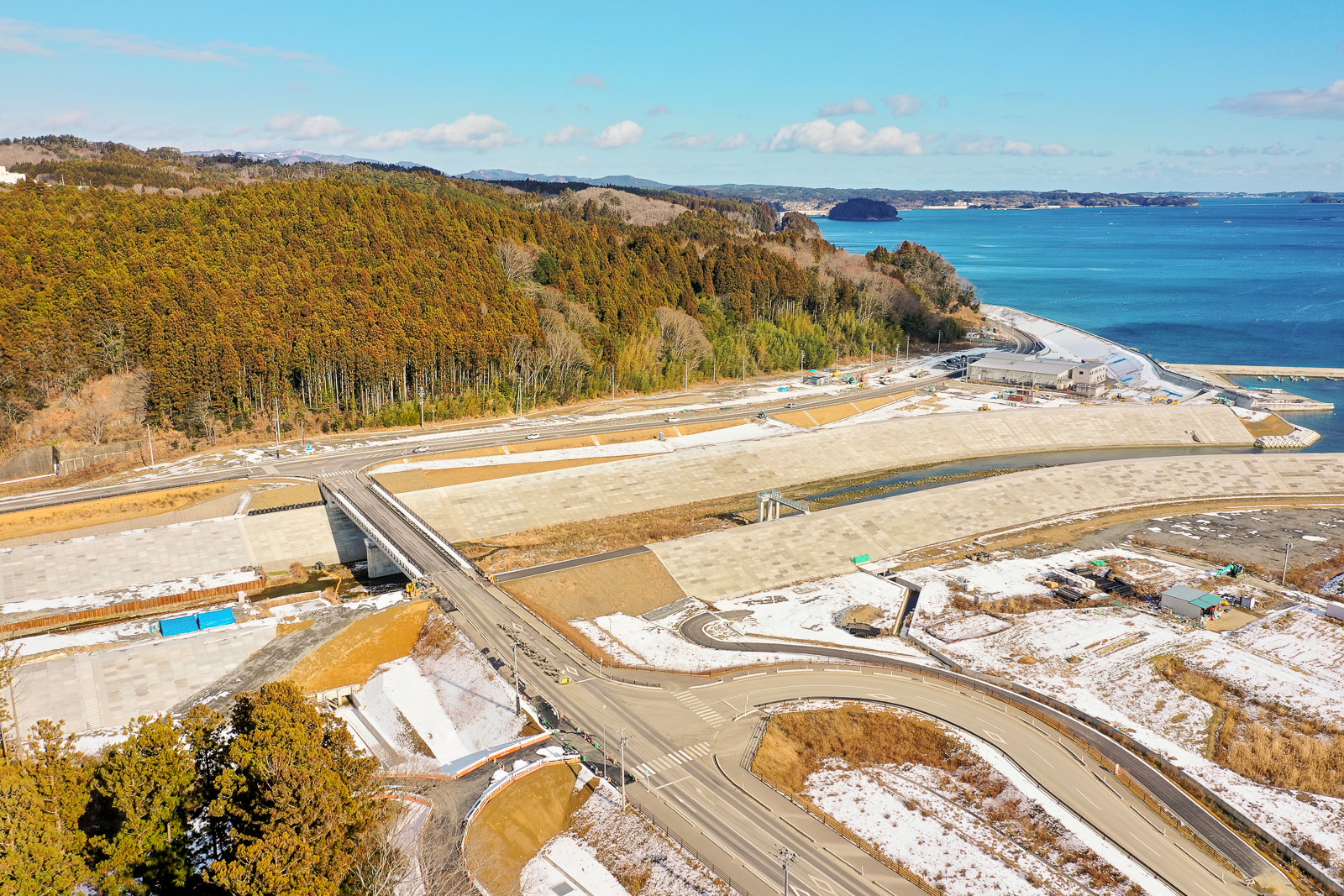 道路網と雪に覆われた地面が広がる海岸地域を上空から撮影した風景で、背景には水辺が広がり、森林に覆われた丘が見られ、橋や建設現場と思われるインフラも点在し、空は晴れて雲が少し浮かぶ冬の海岸地域の写真