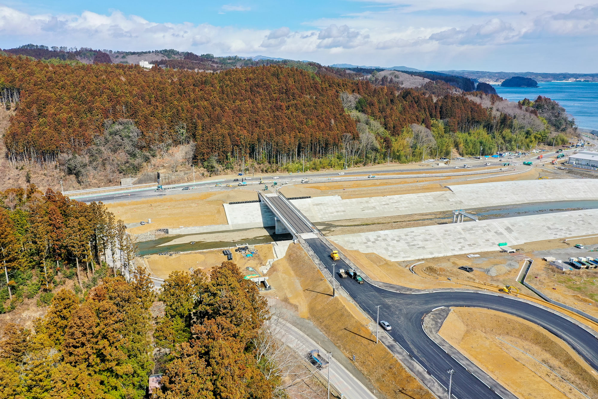 小さな川または運河に架けられた新しい橋を中心とした工事現場を空から撮影した写真で、周囲には森林に覆われた丘や道路、建設機材が点在する写真