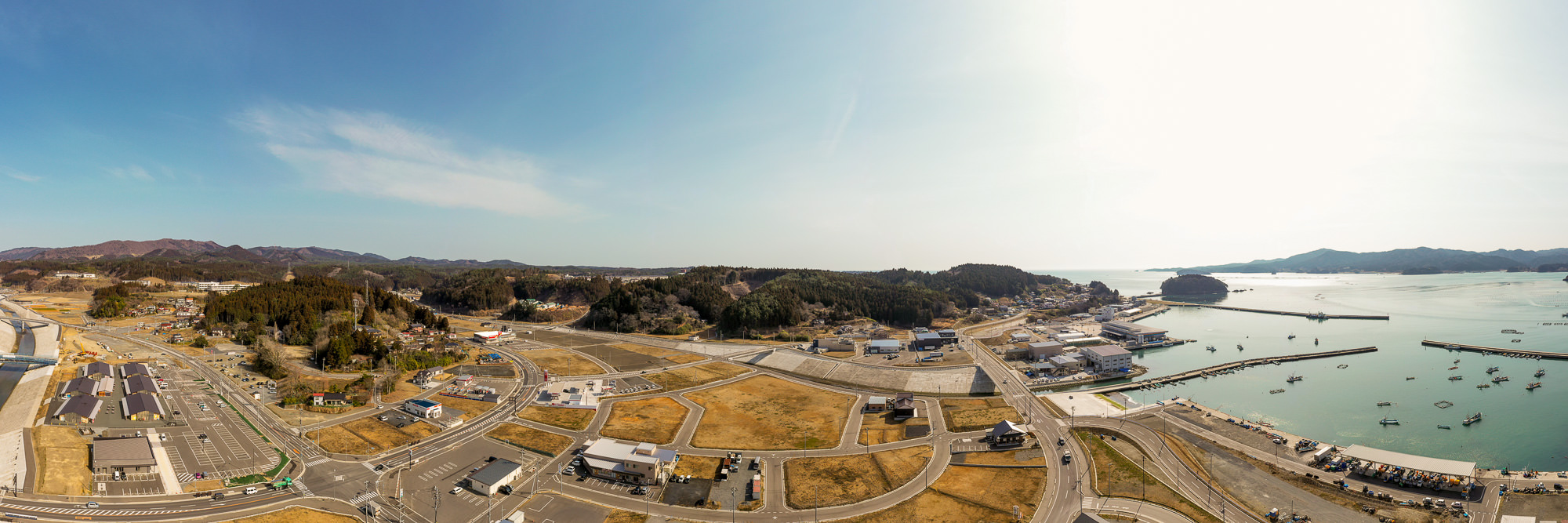 建物や道路が広がる港町を高所から見渡し、周囲には丘と海が広がり、海には複数の船が浮かび、空は雲が少なく晴れている様子を写した海辺の町の写真