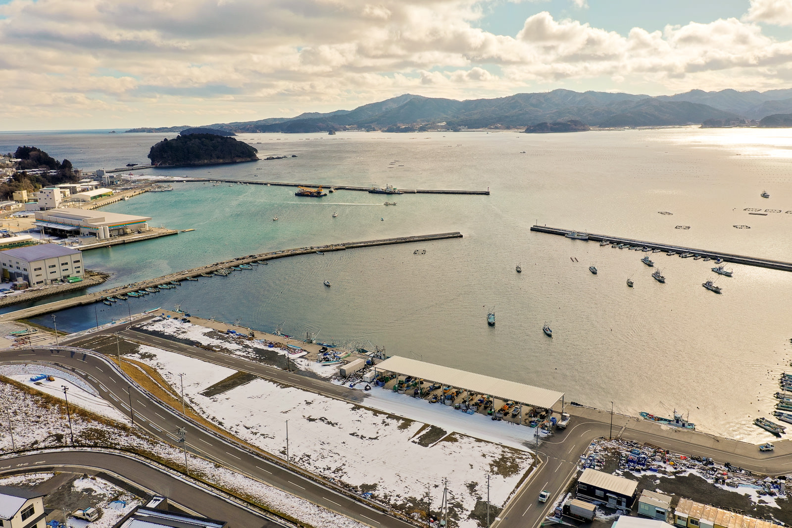海に面した地域を広く見渡す構図で、港には複数の桟橋が伸び、船が水面に浮かび、海岸沿いには建物が並び、手前の地面には雪が積もっており、背景には山々と遠くに島が見え、空には雲が点在する冬の海辺の港の写真