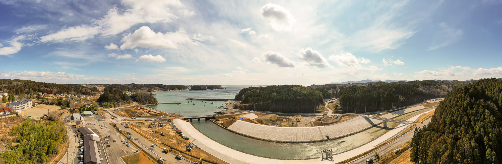 広い空の下、川が海へと流れ込む地点を中心に、周囲には緑に覆われた山々と点在する建物が見られる自然と人の営みが調和した風景の写真