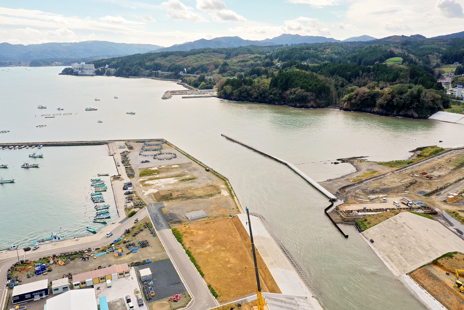 川が海へと流れ込む沿岸地域を空から捉えた風景で、岸辺には複数の船が停泊し、周囲には建設現場や建物が点在し、背景には丘や山々が広がる海辺の町の写真