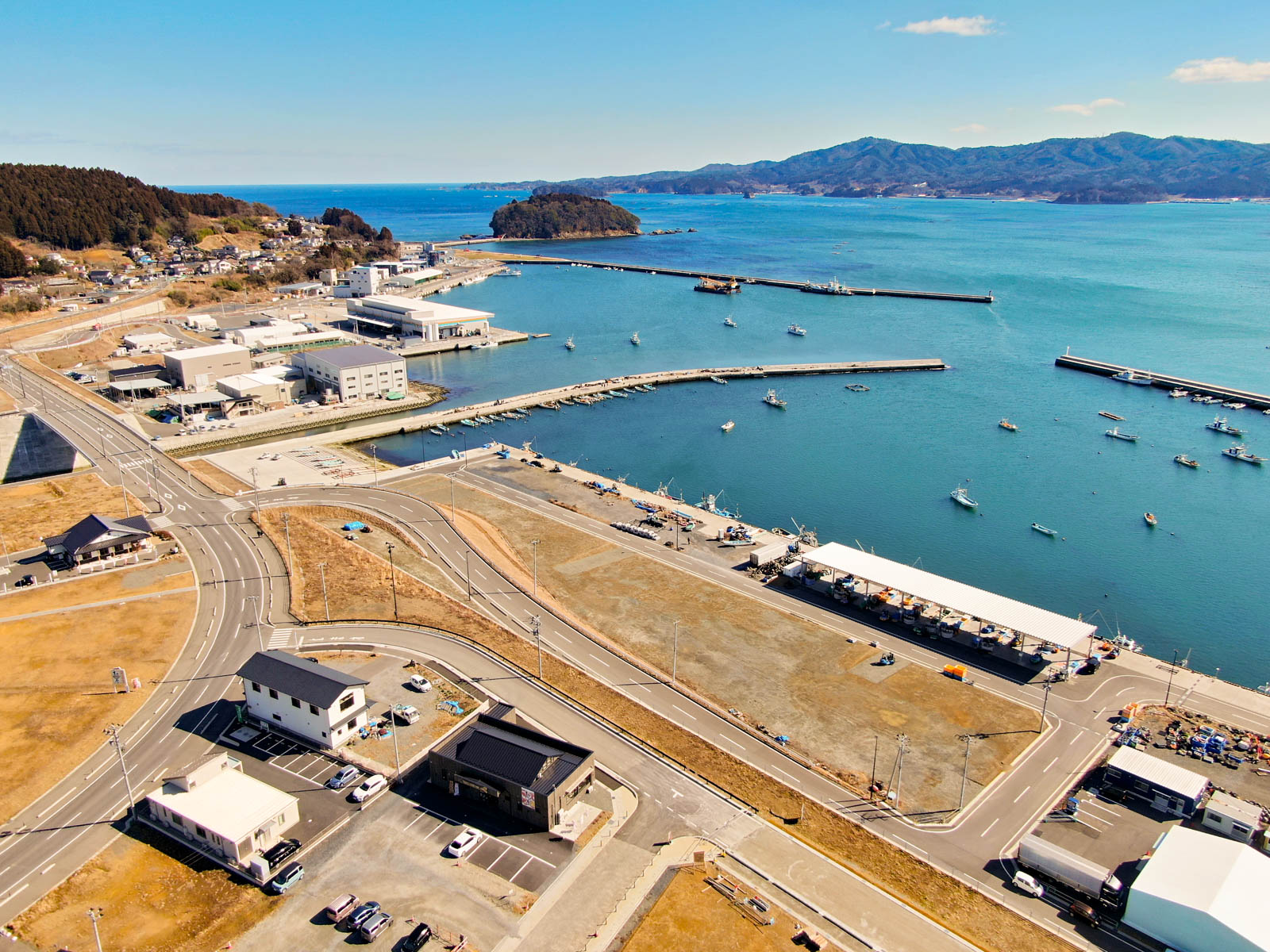 港を中心とした海沿いの町を上空から見下ろした風景が広がっており、港には複数の船が停泊し、いくつもの桟橋が水面に突き出していて、周囲には建物や道路、広場が点在し、背景には丘や山々、さらに海の向こうには島が見え、空にはわずかに雲が浮かぶ晴天の写真