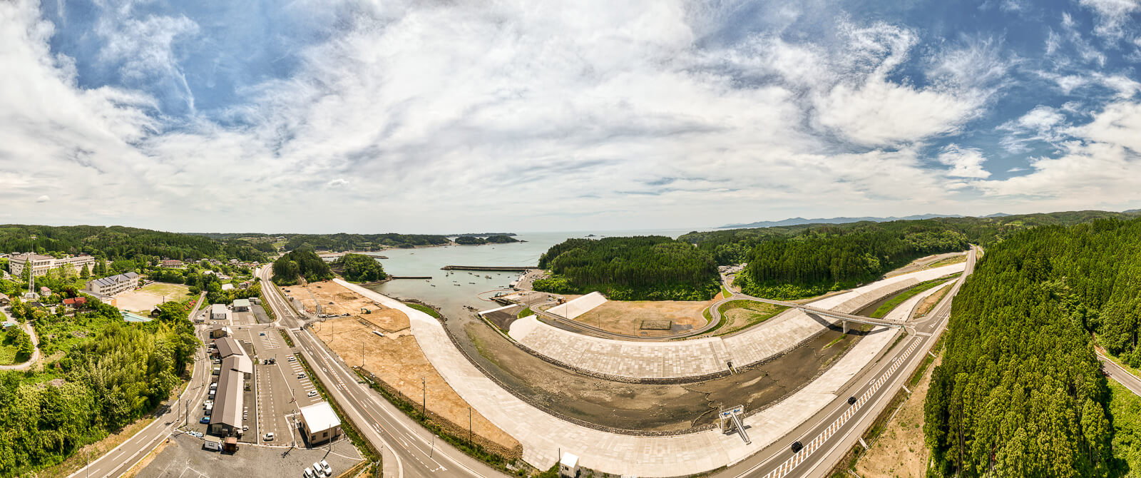 海沿いに広がる防波堤がU字型に建設されていて、その周囲に道路や林、建物や駐車場が点在し、遠くに小さな島々が見える昼間の風景を高台から定点カメラで撮影した写真