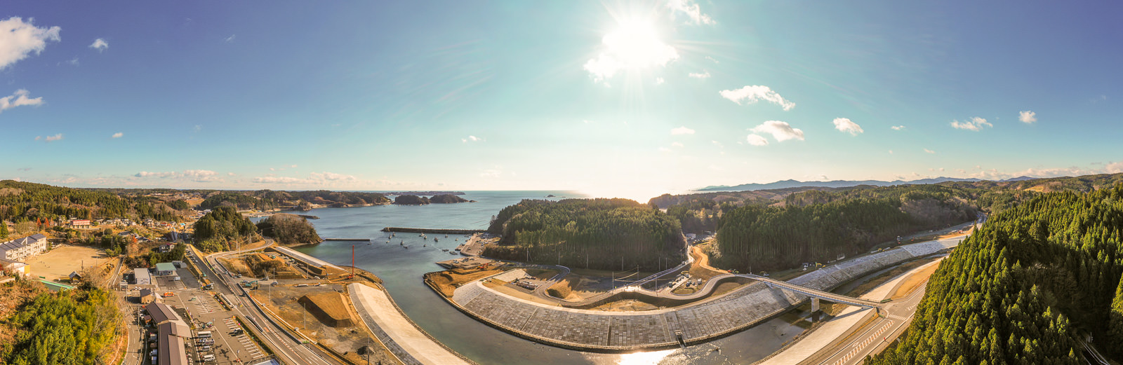 川が海へと流れ込む海岸地域を広く見渡した風景で、川沿いにはダムや堤防のような構造物があり、周囲は森林に覆われた丘に囲まれ、いくつかの建物が点在し、空は晴れて雲が少し浮かび、水面には太陽の光が反射し、下部には道路やインフラが整備された自然と人の営みが共存する海岸地域の写真