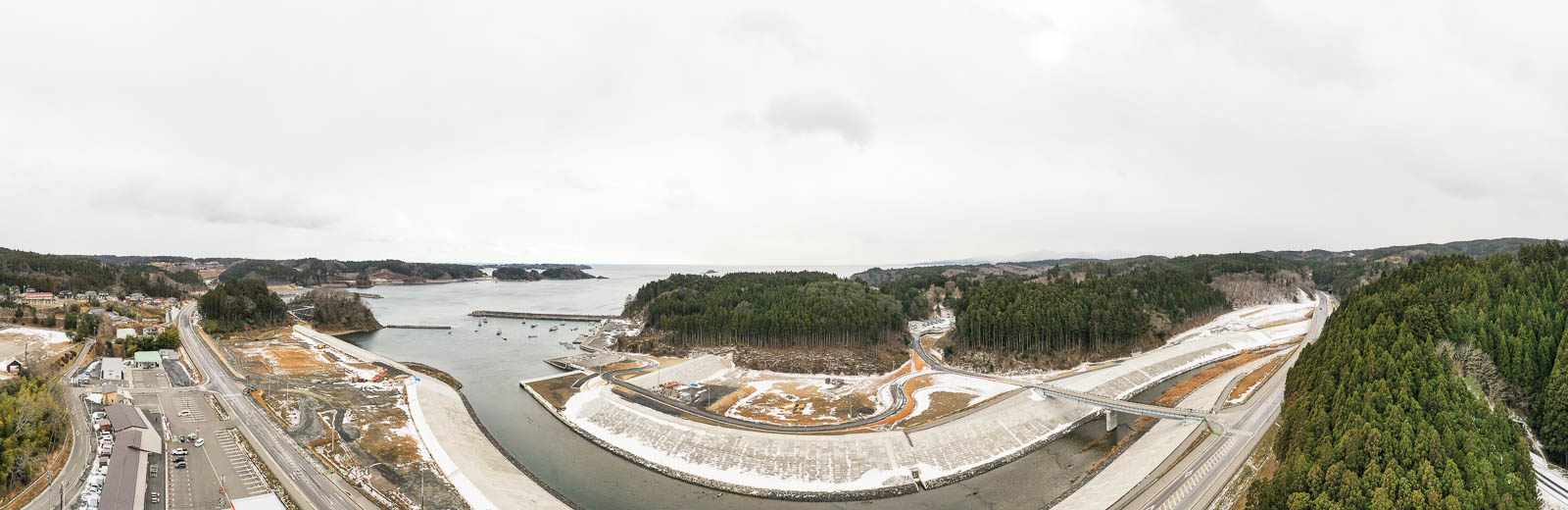 川が湾に流れ込む地点に大規模な護岸と橋が架けられ、両岸は山林に囲まれ、海の先にいくつかの島が見える再開発中の港湾の昼間の風景を高台から定点カメラで撮影した写真